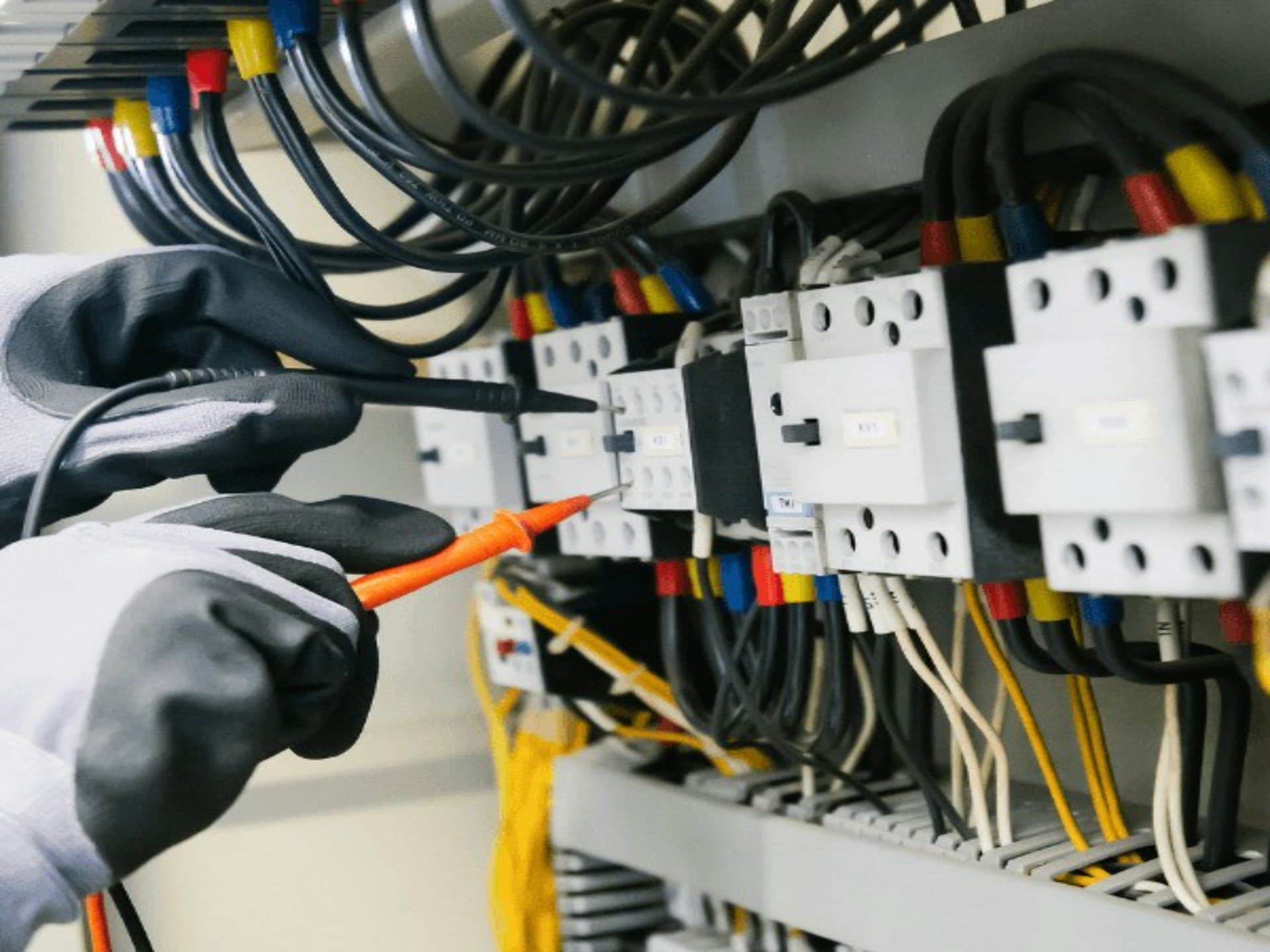 Gloved electrician testing wiring on an industrial control panel with a multimeter probe