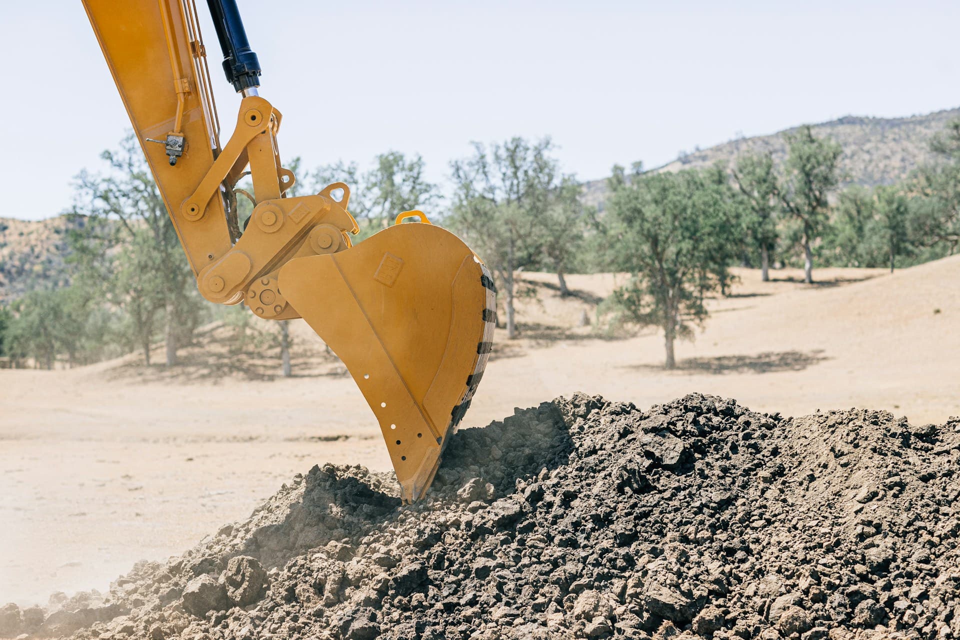 Excavator bucket lifting a load of dark soil at a dry rural site