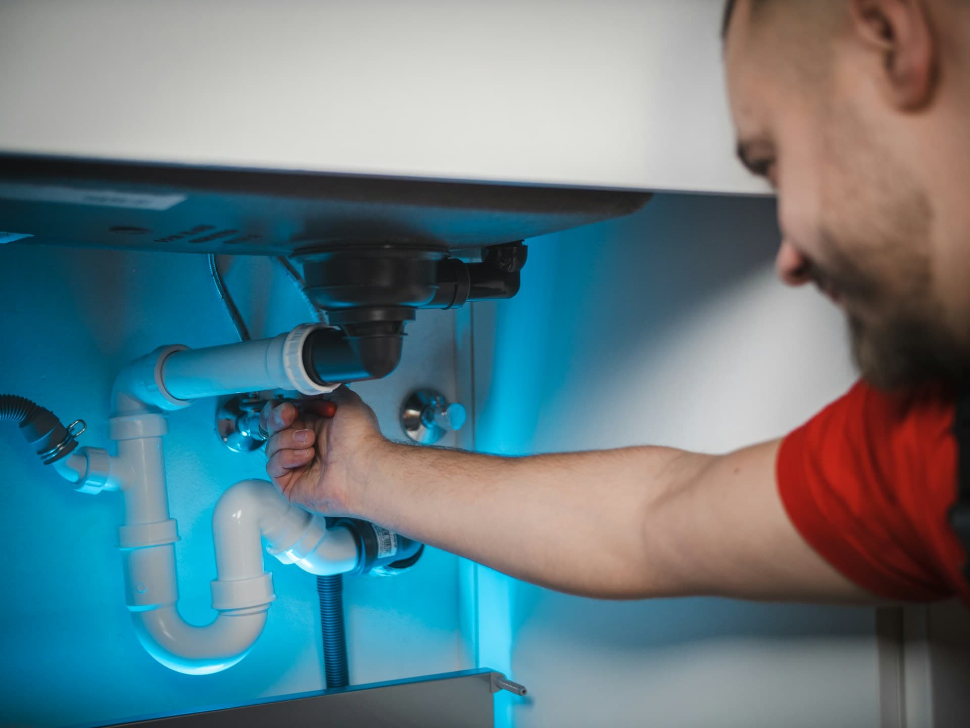 Plumber in a red shirt tightening pipework under a sink drain trap
