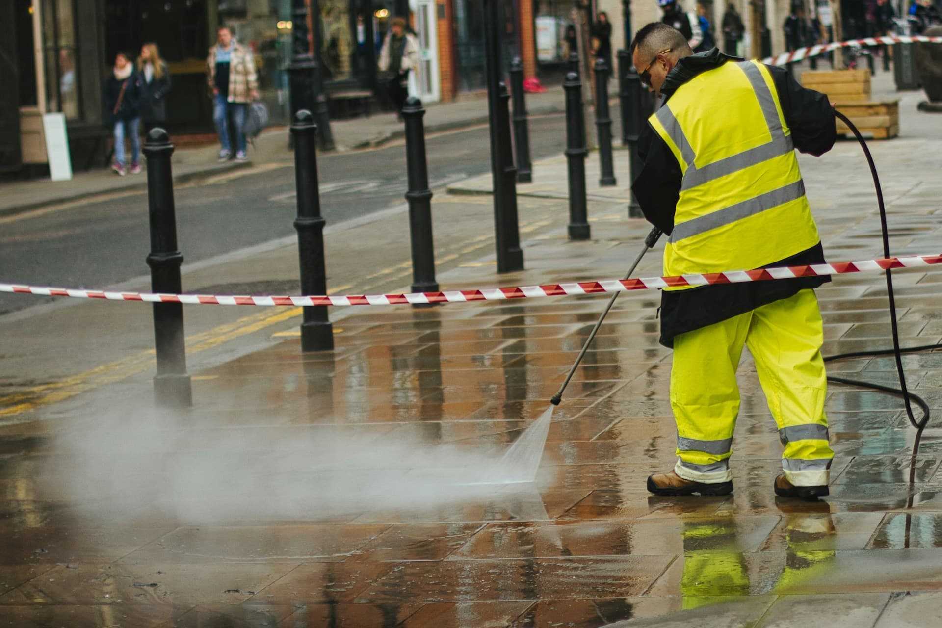 Worker in hi-vis waterproofs pressure-washing a wet paved street behind hazard tape