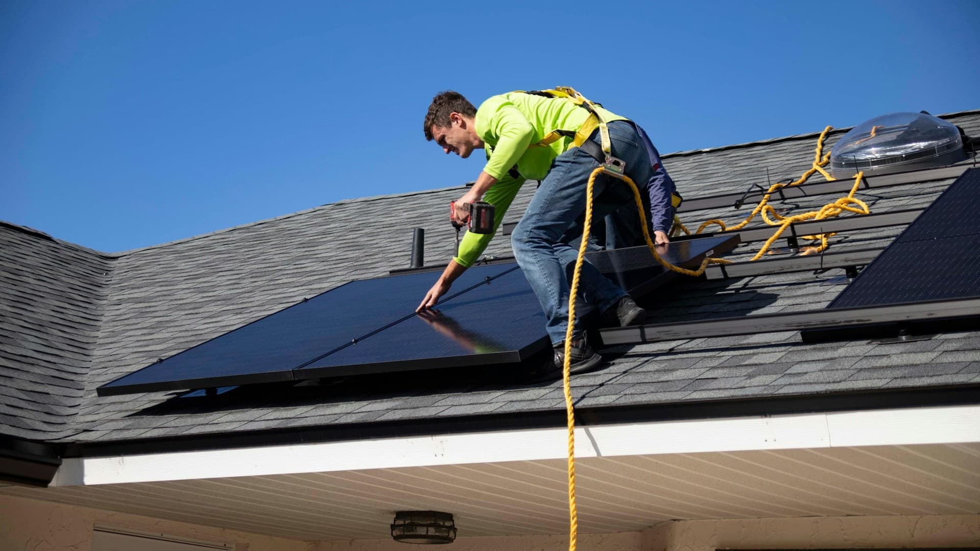 Worker in hi-vis shirt and harness drilling a solar panel on a shingled roof