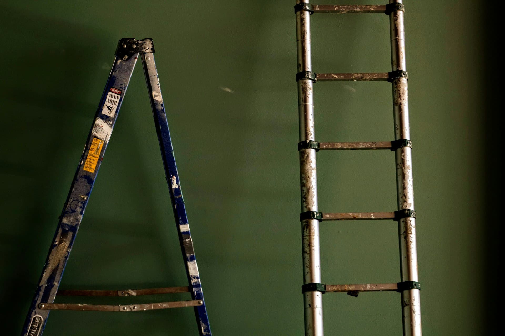 Paint-spattered aluminium step ladder and extension ladder against a green wall