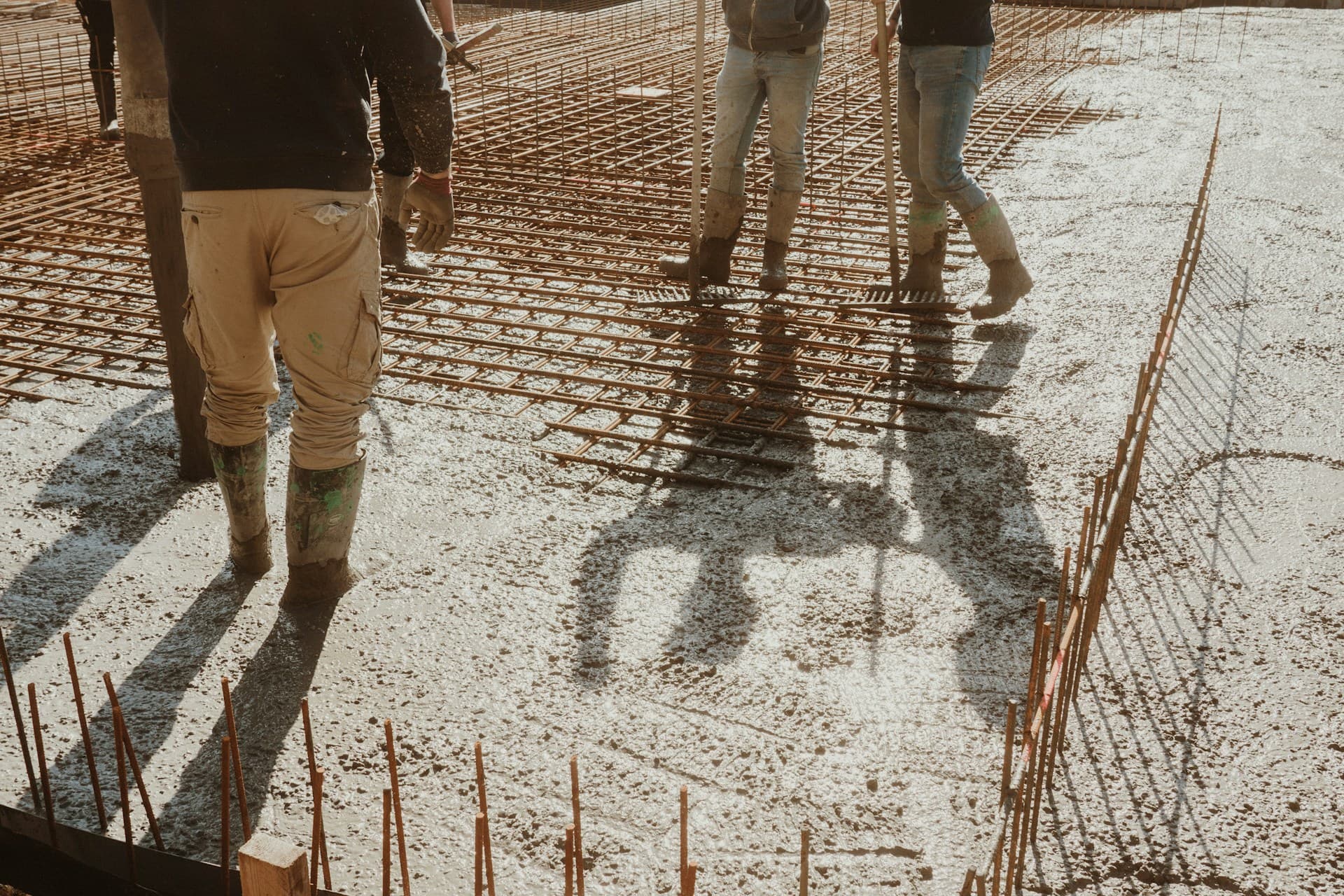 Workers in boots raking wet concrete over steel mesh reinforcement in bright sun