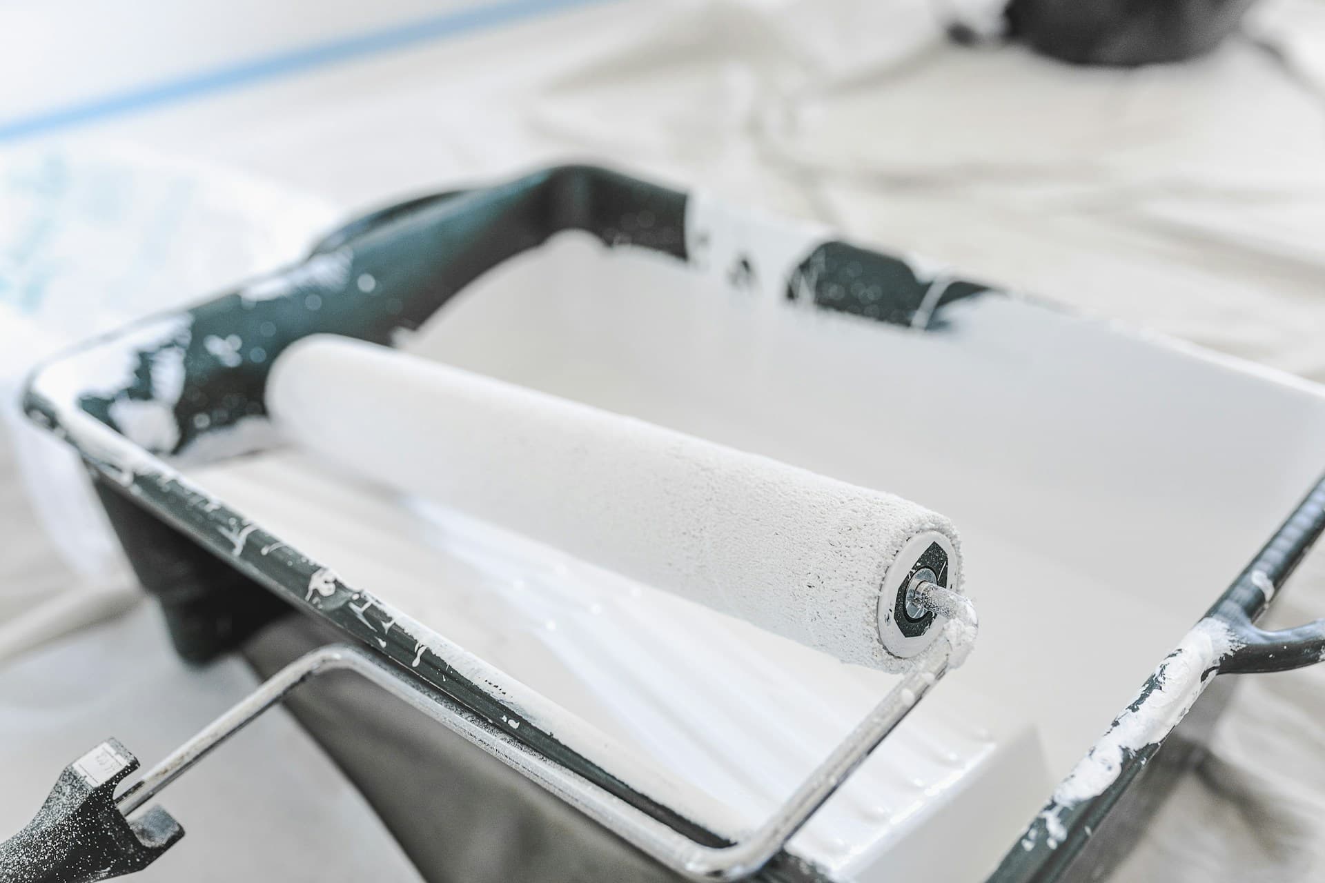 Paint roller loaded with white paint resting in a tray on drop sheets
