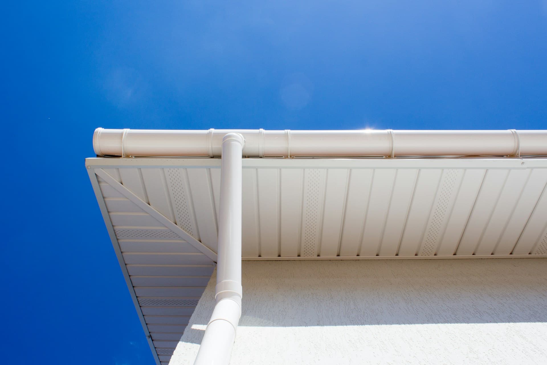 White roof gutter and downpipe along fascia against a clear blue sky
