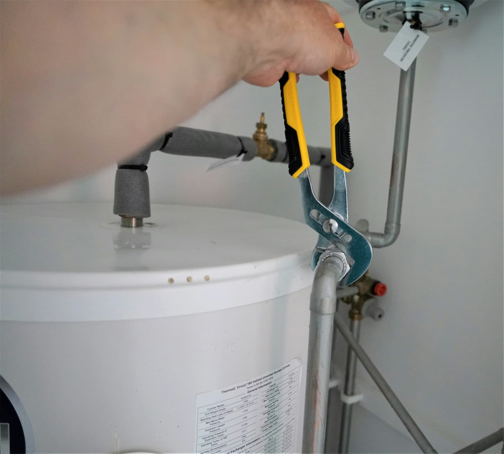 Hand using yellow multi-grip pliers on pipework above a hot water cylinder