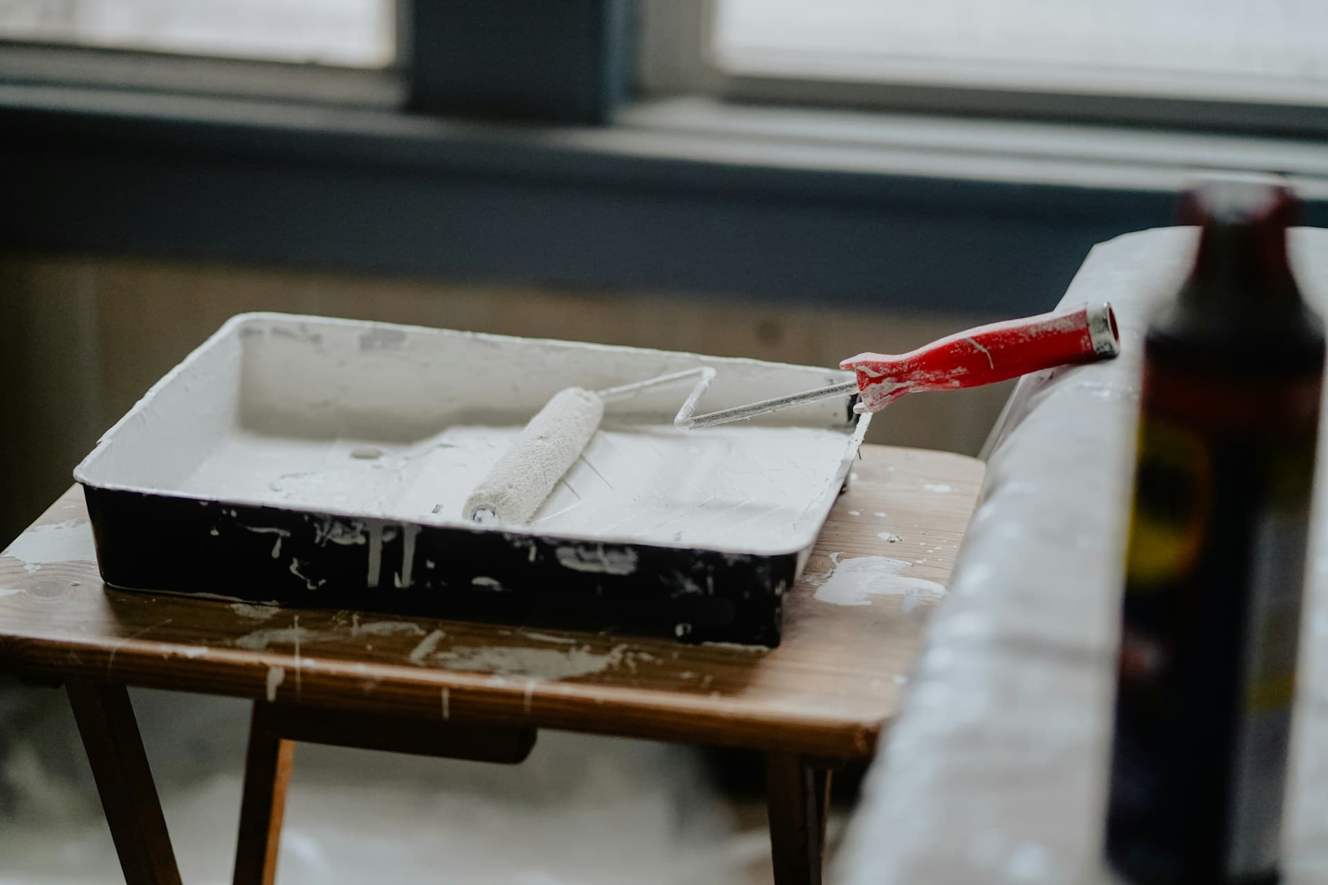 Paint roller and white-filled tray on a paint-spattered wooden table by a window