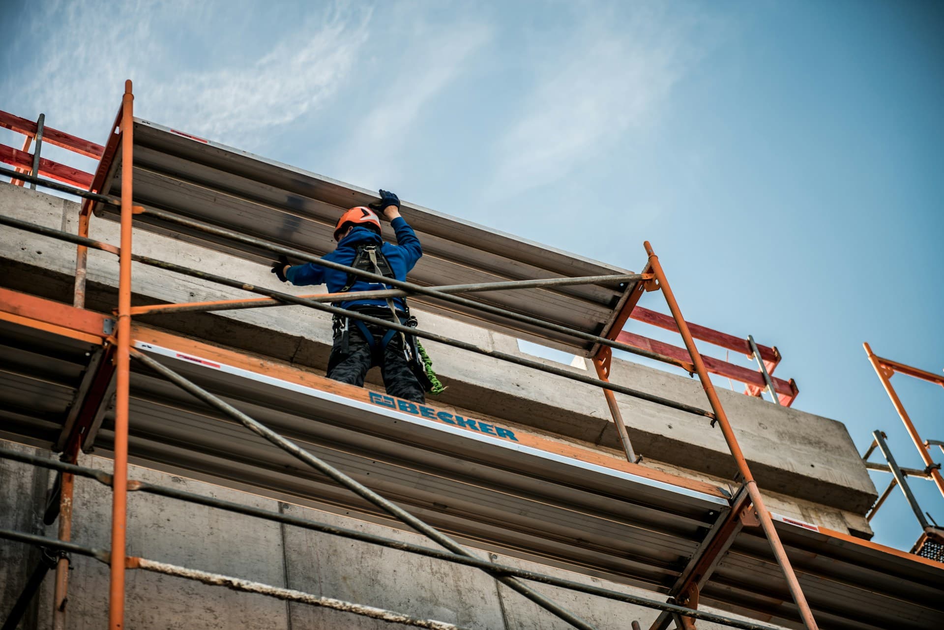 Worker in helmet and harness on scaffolding handling a metal panel against the sky