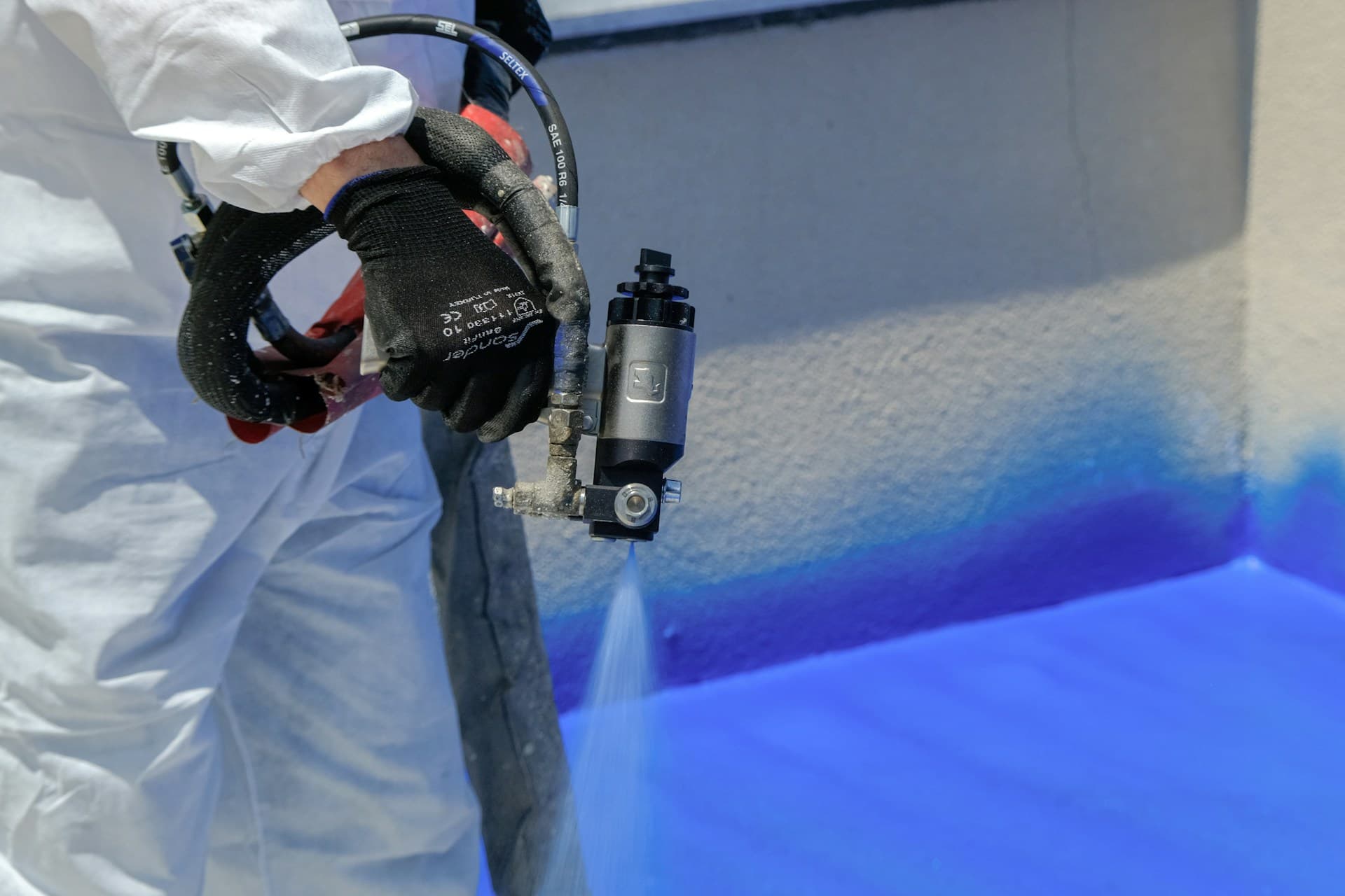 Worker in white coveralls and gloves applying blue spray coating with a sprayer