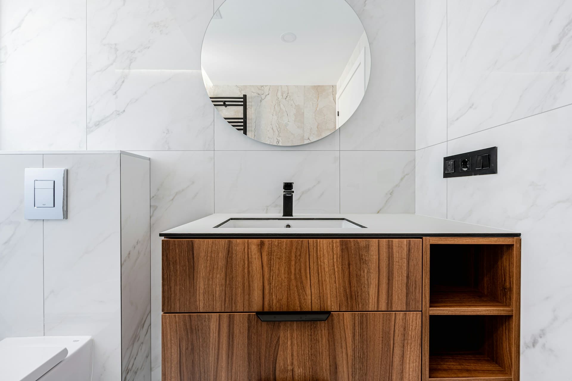 Marble-tiled bathroom wall with round mirror, timber vanity and black tapware